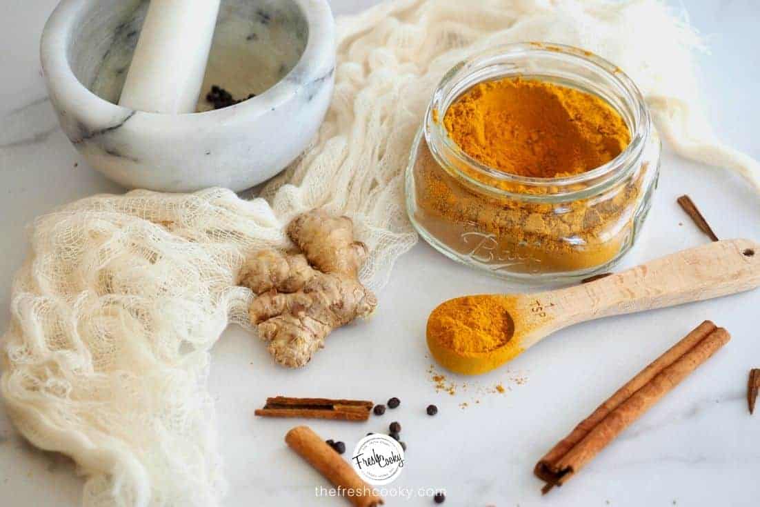 A mini wide mouth mason jar filled with Turmeric Tea Spice Mix with a wooden spoon with the tea in it, ginger, cinnamon spice and a mortar and pestle.