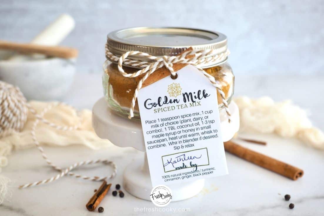 Jar of Golden Milk Spice Mix on a mini pedestal with a gift label tied around it and cinnamon sticks behind.