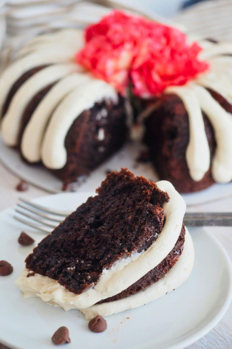 Rich chocolate cake slice on plate with full bundt cake in background with fingers of cream cheese frosting. 