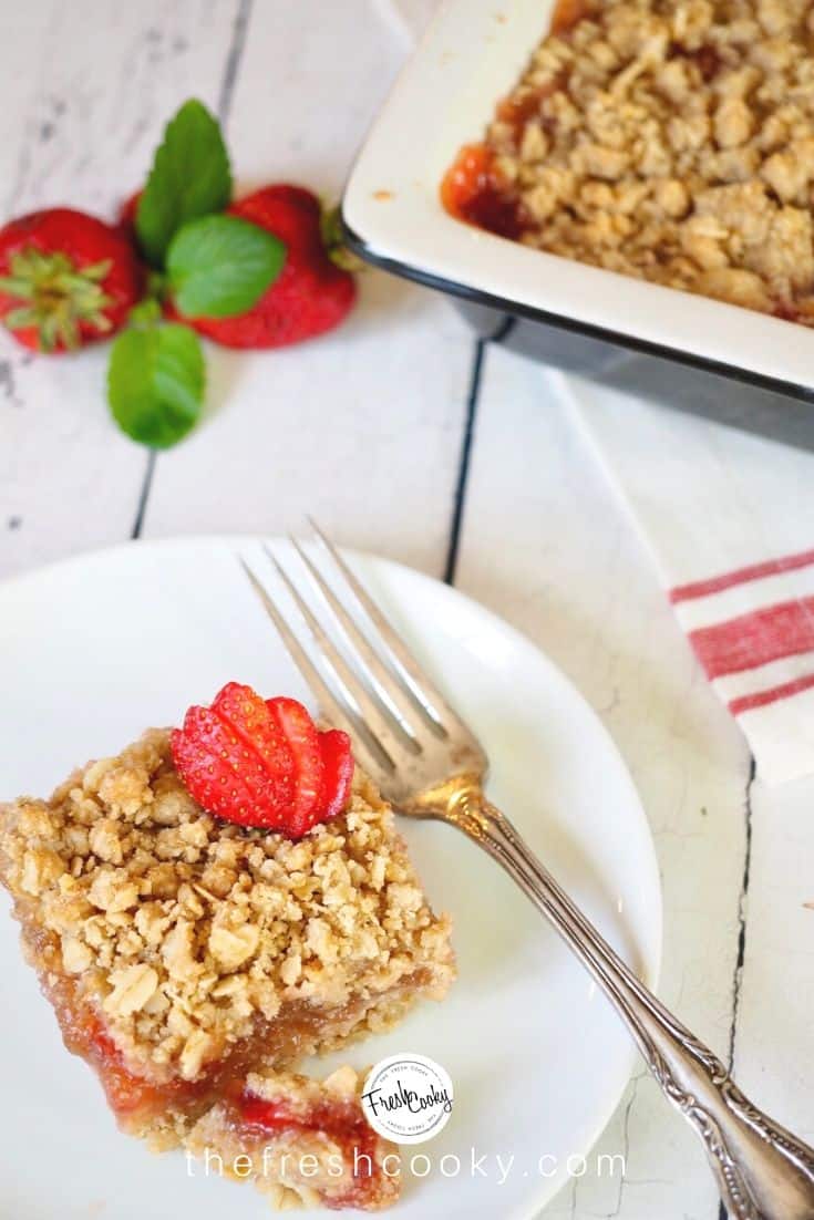 white plate with silver fork and square of rhubarb strawberry crumble bar with sliced strawberry on top and pan of rhubarb crumble in background. 