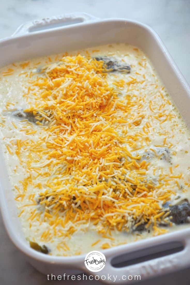 A casserole dish filled with cheese stuffed hatch chile peppers, with an egg, flour and milk mixture poured on top, topped with grated cheese. | thefreshcooky.com 