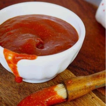 white bowl with barbecue sauce inside, and a wooden handled basting brush with boar bristles with barbecue sauce on a cutting board.