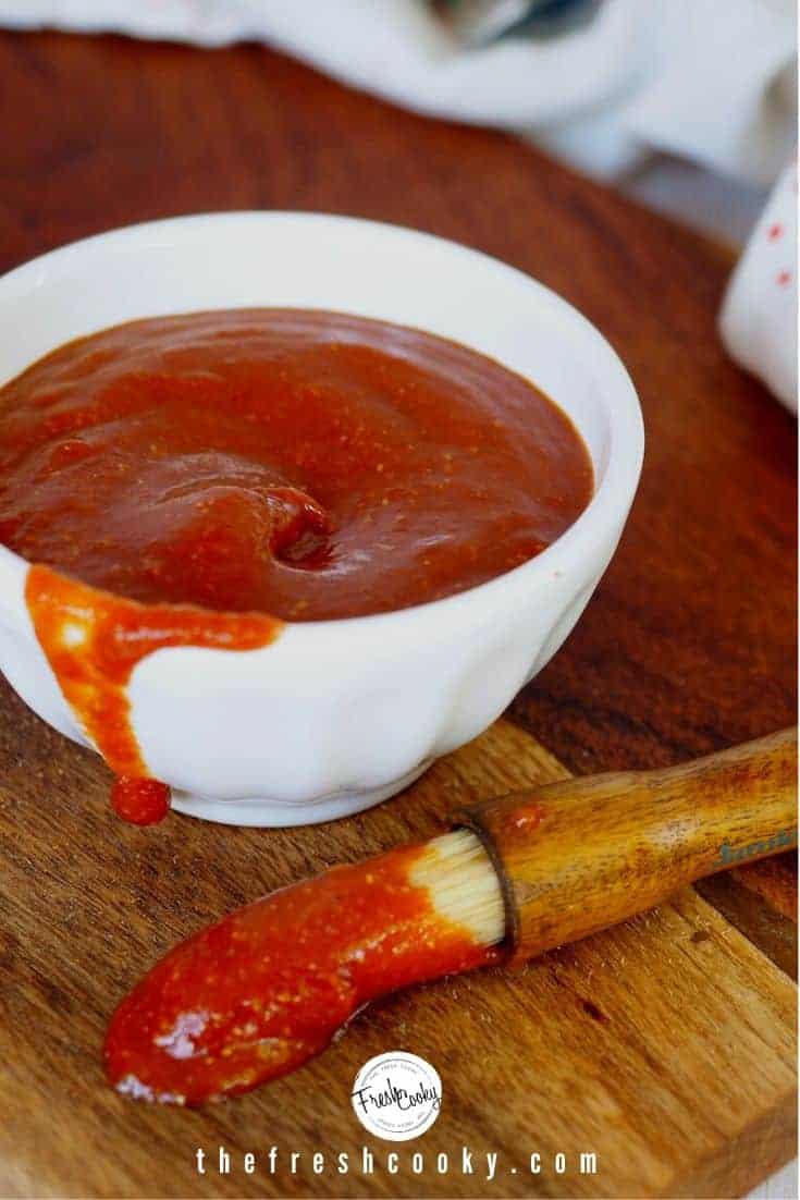 White bowl with barbecue sauce inside, and a wooden handled basting brush with boar bristles with barbecue sauce on a cutting board.