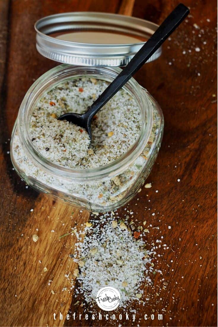 Small glass mason jar with lid filled with Copycat Carrabba's Grill Seasoning, some spilled on cutting board with black spoon in jar.