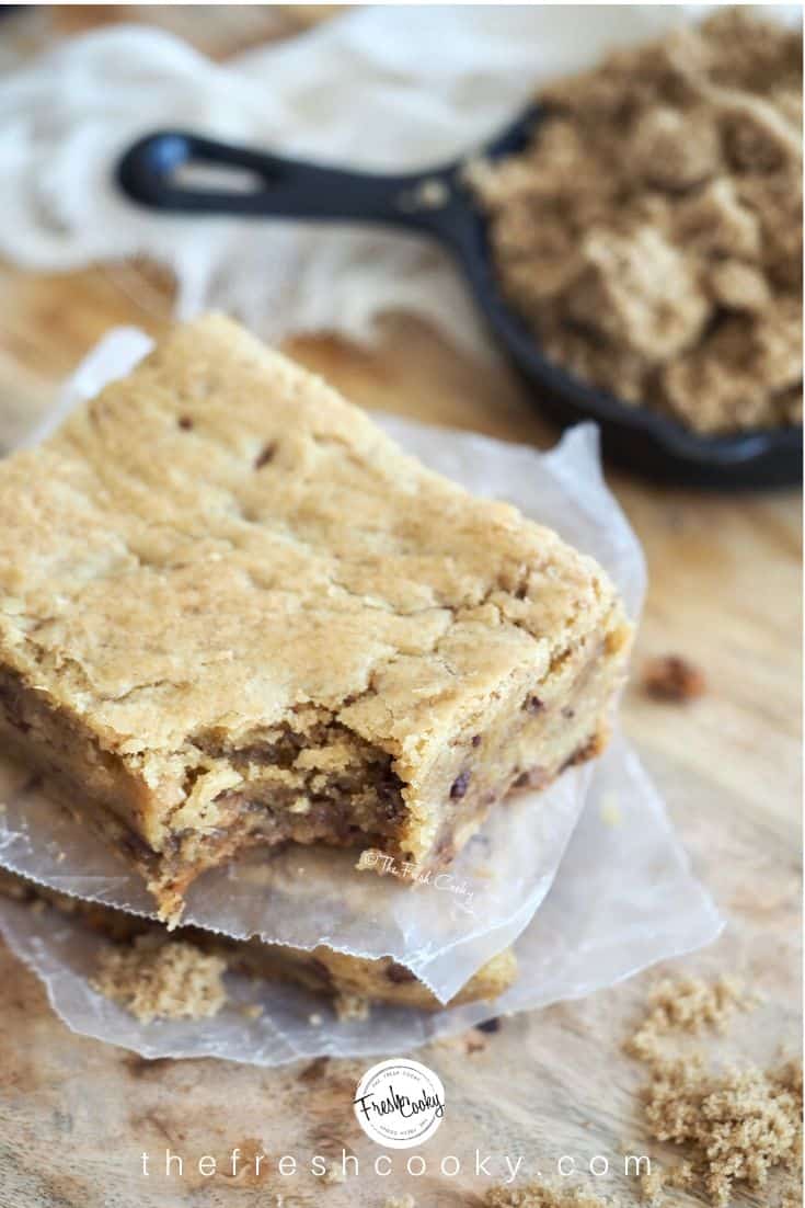 Chewy blondie bar with wax paper between, with bite taken out to reveal gooey insides, background brown sugar in mini cast iron pan.