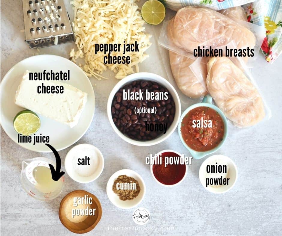 ingredient shot for crack chicken with neufchatel cheese on white plate with a lime, grater with shredded pepper jack cheese, 4 chicken breasts, a bowl of salsa, black beans in white bowl, and onion powder, chili powder, salt, cumin and garlic powder in small white bowls with a mini measuring cup with lime juice.