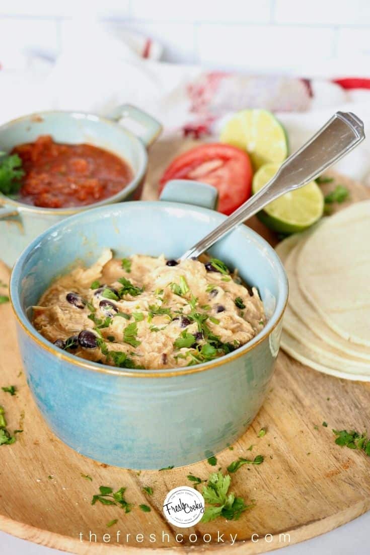 blue ceramic bowl filled with shredded crack chicken on a wooden plate with a bowl of salsa, limes, tomatoes and corn tortillas