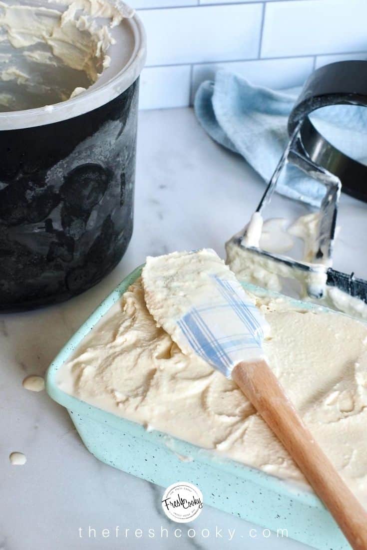 Ice cream freezer in background with scraper and in foreground, an aqua loaf pan filled with vanilla ice cream and a blue and white plaid spatula used to smooth it on top.