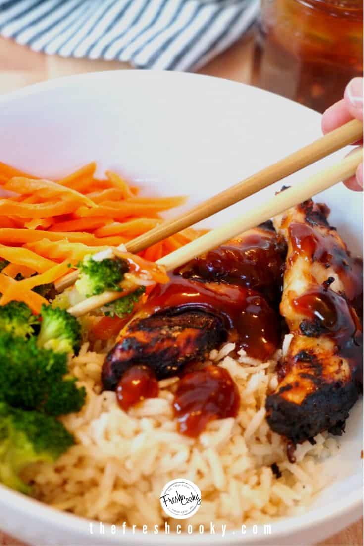 Chopsticks picking up broccoli and teriyaki chicken from large white bowl with brown rice, grilled chicken, shredded carrots, steamed broccoli drizzled with teriyaki sauce.