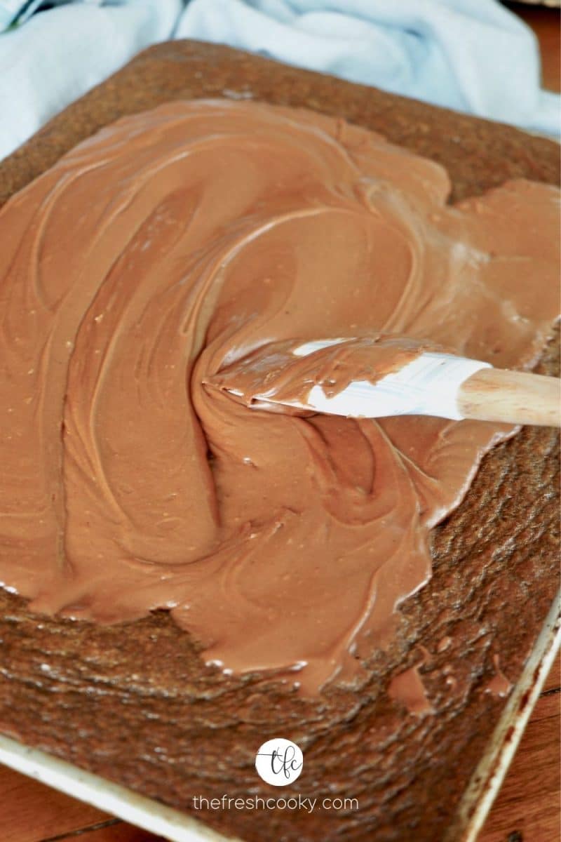 A spatula smoothing on cooked fudge frosting onto the top of a Texas Chocolate Sheet Cake. 