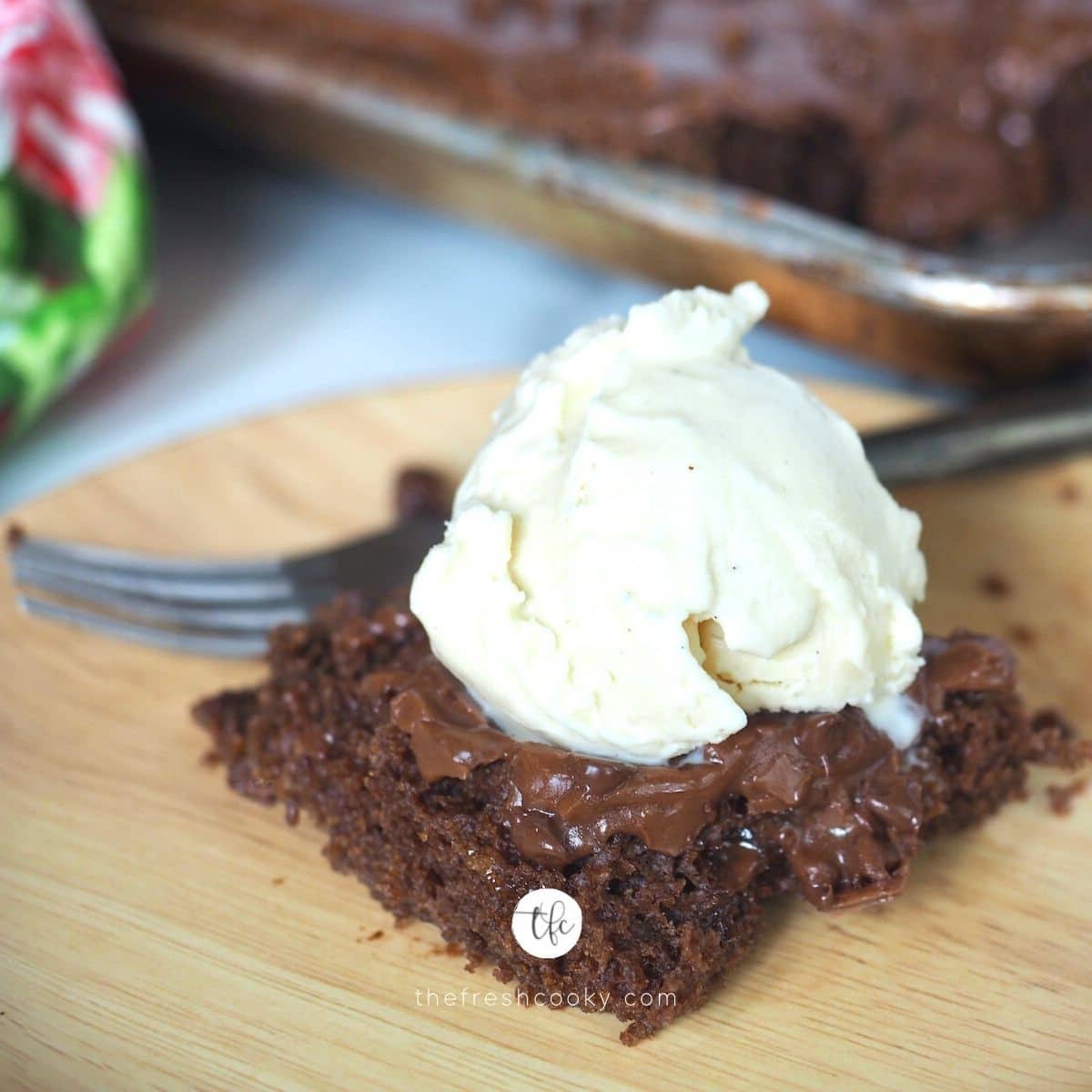 Square image of slice of chewy, gooey Texas sheet cake topped with a scoop of vanilla ice cream, full sheetcake in background.