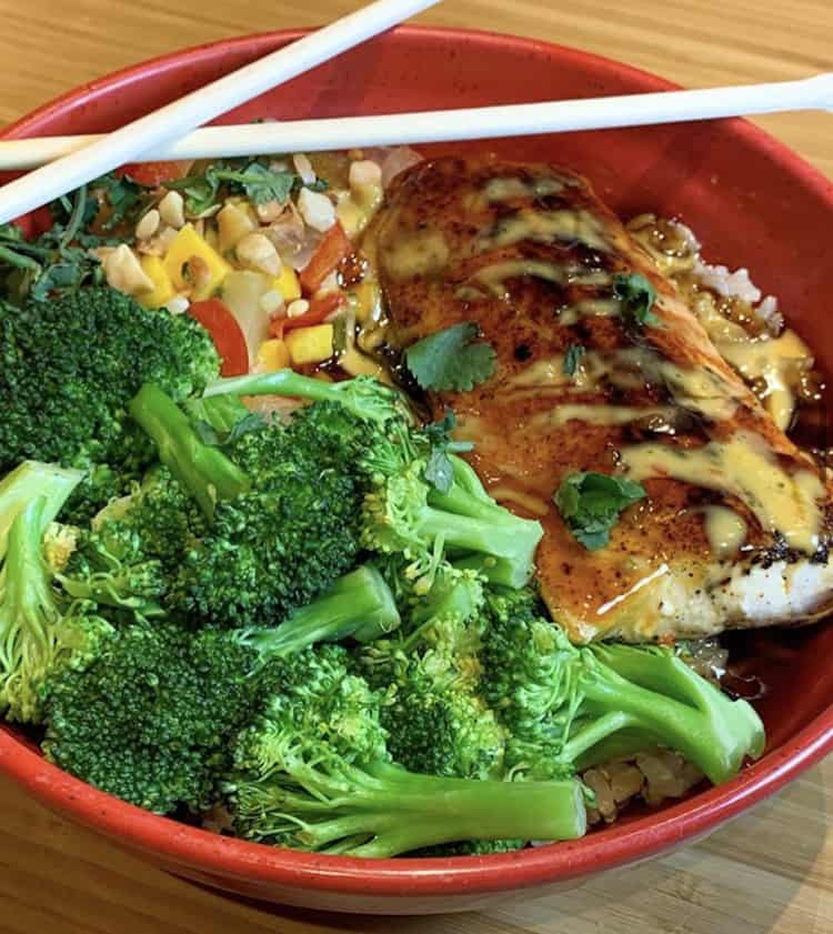 Red bowl filled with brown rice, steamed broccoli a corn salsa, mahi-mahi grilled fish with Teriyaki Sauce and chopsticks crossed atop the bowl.