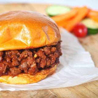 Sloppy Joe sandwich on top of white parchment paper on a wooden charger with bright carrots, cucumbers and tomatoes in background