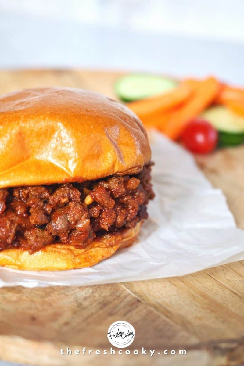 Sloppy Joe sandwich on top of white parchment paper on a wooden charger with bright carrots, cucumbers and tomatoes in background