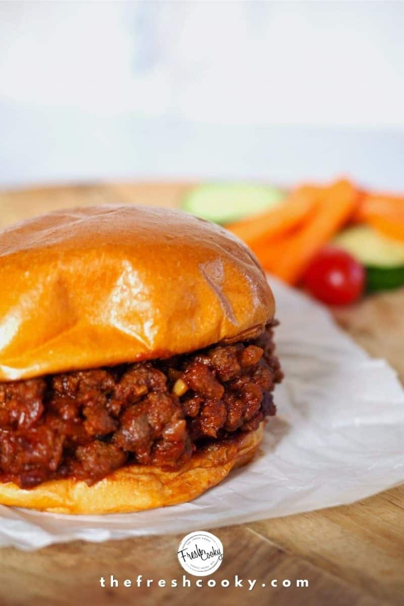 Sloppy Joe sandwich on top of white parchment paper on a wooden charger with bright carrots, cucumbers and tomatoes in background