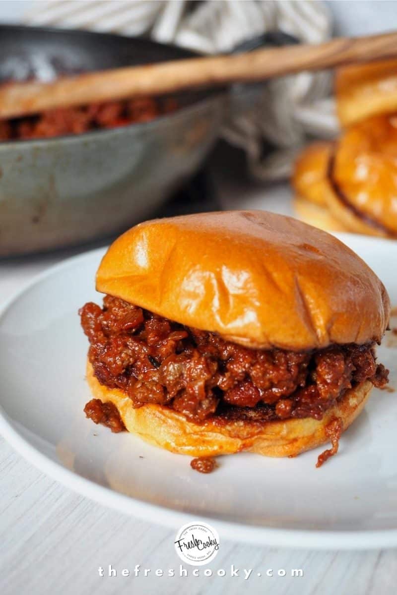 Sloppy Joe Burger ona white plate with brioche buns in background along with a pan with sloppy joe mixture and wooden spoon