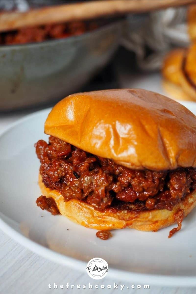 White plate with sloppy joe burger on it stuffed with sloppy joe filling with skillet in background with wooden spoon resting inside