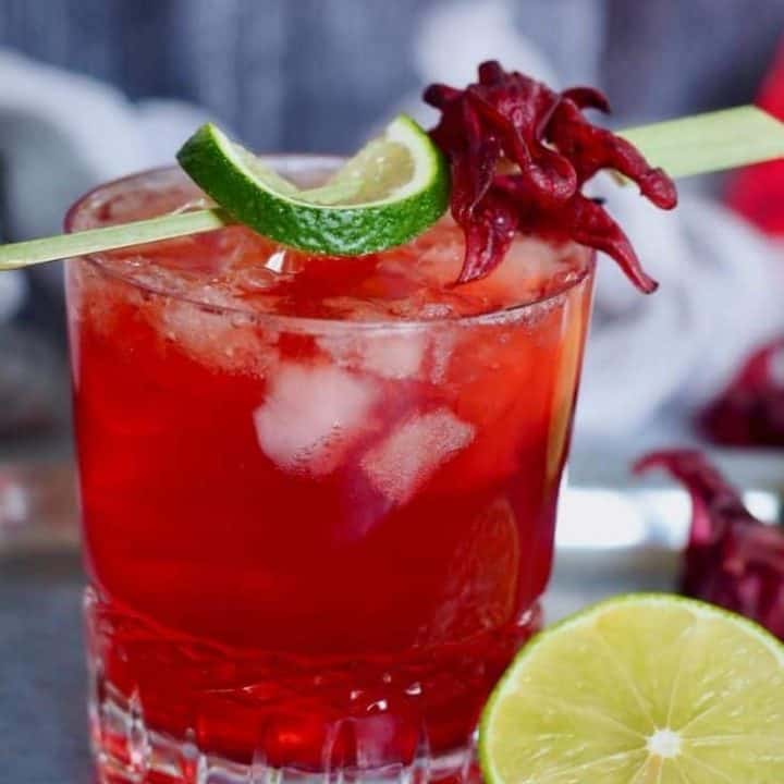 crystal glass filled with red liquid, hibiscus ginger beer, dark rum and lime. with a wedge of lime. Garnished with a bamboo stick, lime and hibiscus flower