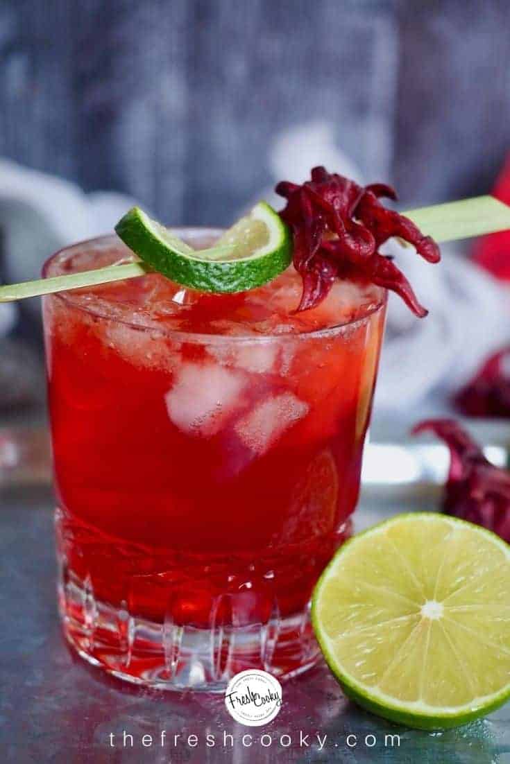 Image of pretty crystal glass filled with bright red cocktail, a hibiscus dark and stormy rum cocktail. Lime half near drink, drink garnish with lime wedge and dried hibiscus flower. 