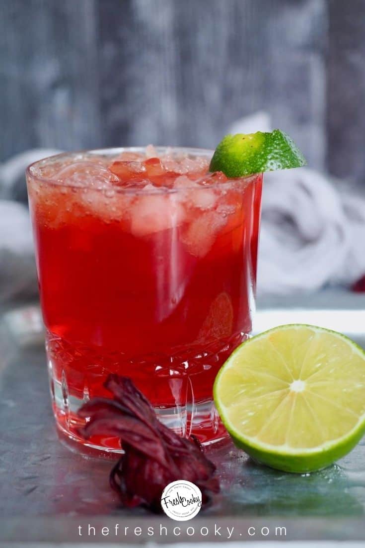 Crystal glass filled with bright red cocktail and crushed ice with lime wedge and half of a lime leaning against the side of the glass.