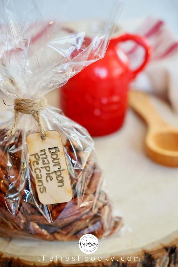 Cello bag filled with maple bourbon pecans tied with twine with a wooden tag handwritten Bourbon Maple Pecans sitting on sliced wood piece with red syrup jar and wooden tablespoon in background.