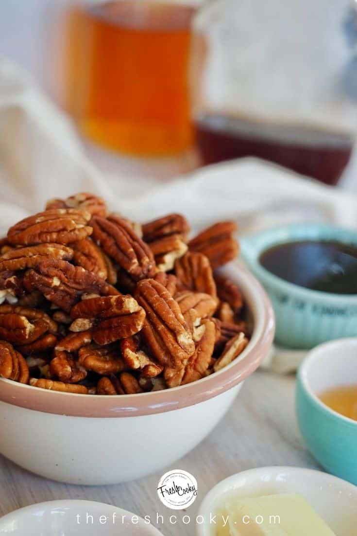 close-up shot of raw pecan halves in small bowl, with maple syrup and bourbon in background.