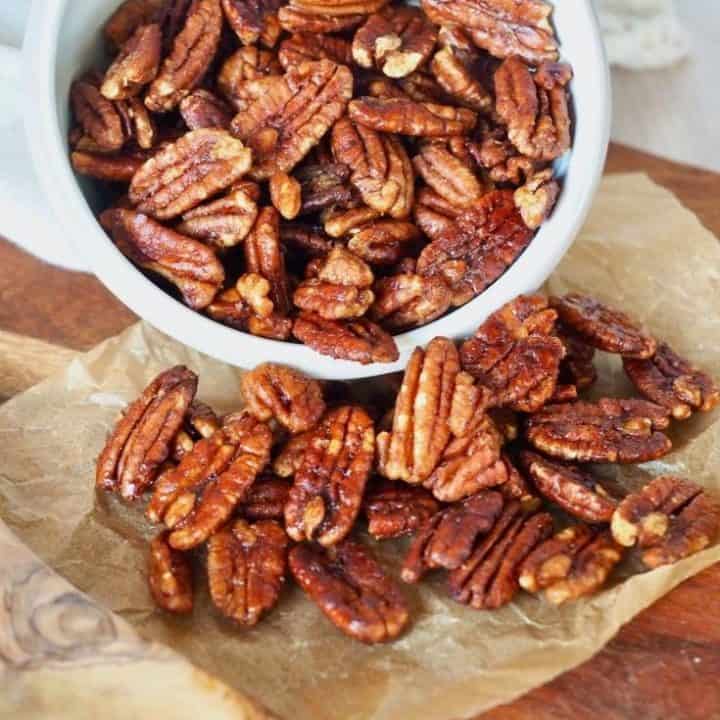 small white bowl tipped, with maple bourbon pecans spilling out onto piece of parchment on wooden cutting board