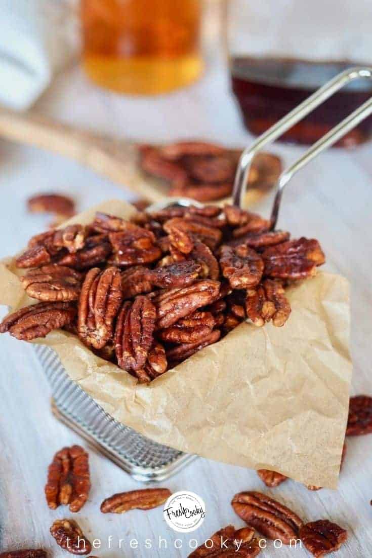 Small wire fry basket lined with parchment paper, holding maple bourbon pecans with wooden spoon, maple syrup and bourbon jars in background. 