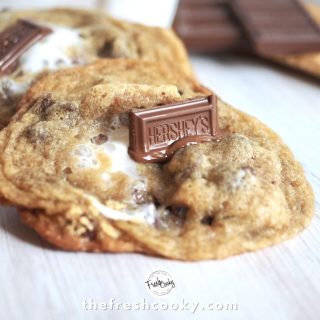 Two large stuff s'more chocolate chip cookies with Hershey bar piece melting on top, graham crackers, marshmallows and chocolate bar in background.
