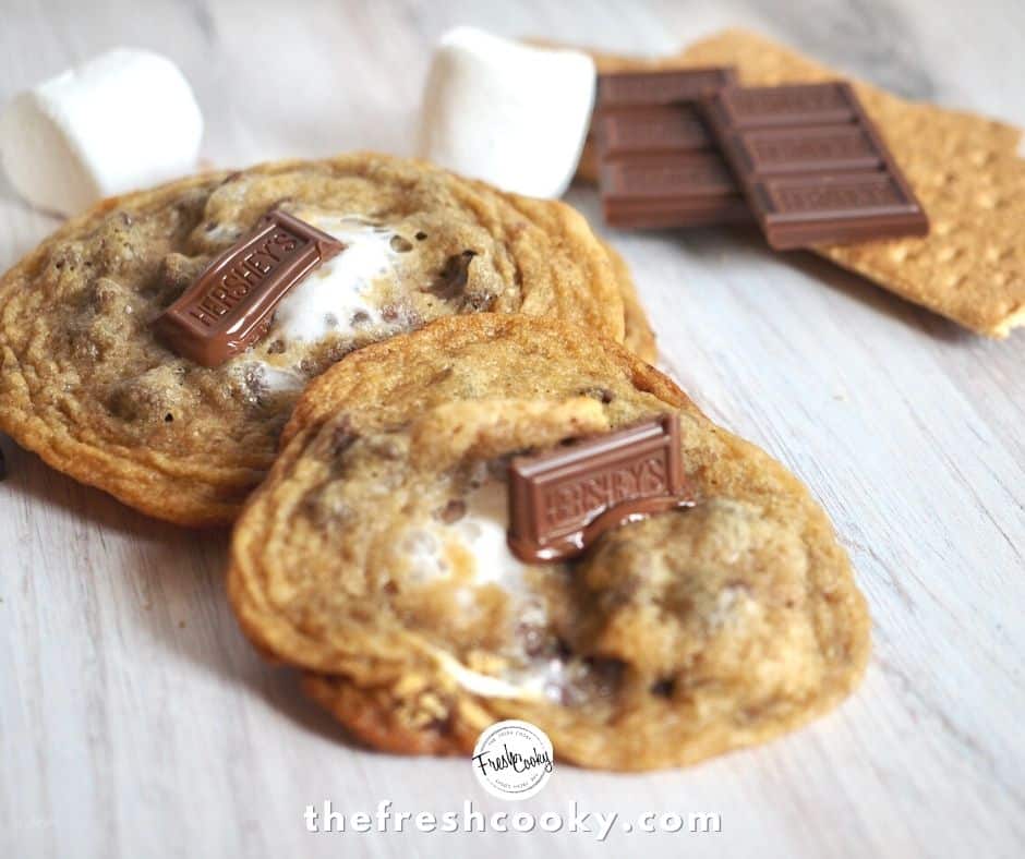 two s'more stuffed cookies sitting on wooden surface with marshmallows, chocolate pieces and graham crackers in background.