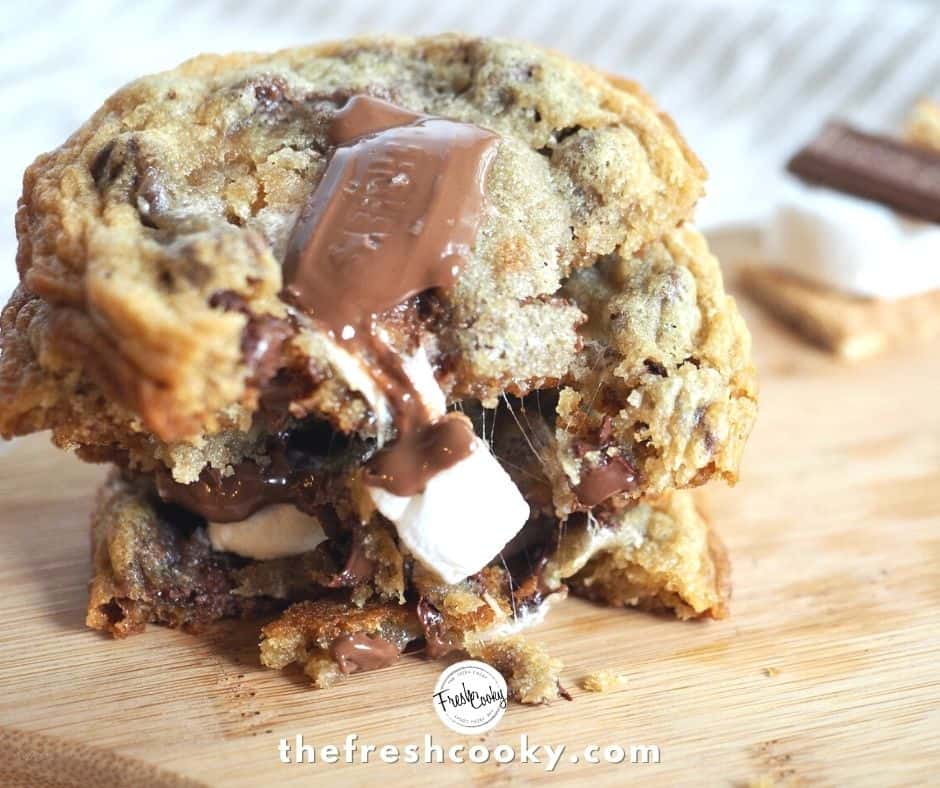 stack of cookies broken in half with gooey marshmallow and chocolate oozing down the sides, sitting on cutting board with striped tea towel in background and small stack of mini s'more