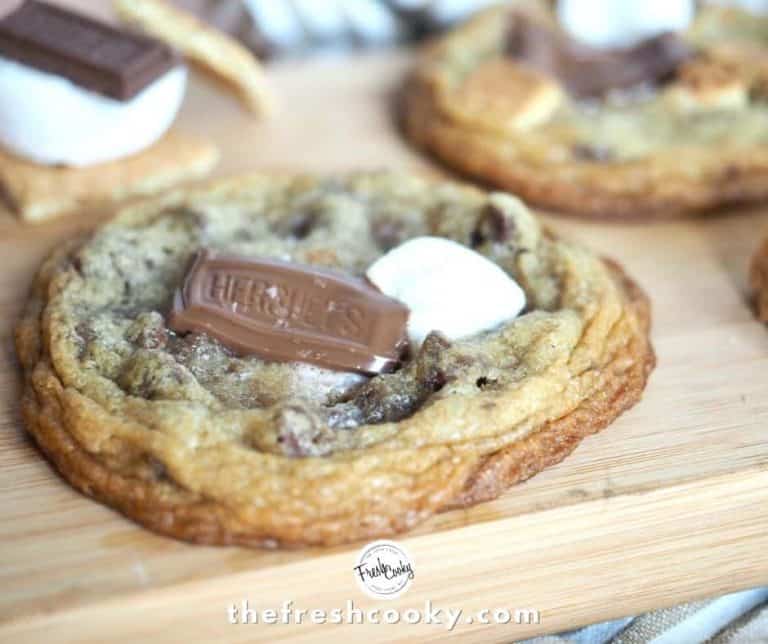 S'more stuffed chocolate chip cookie sitting on cutting board with striped tea towel in background and mini s'more stacks sitting on cutting board.