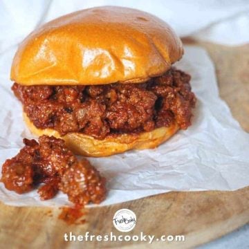 Image of a sloppy joe burger on white parchment paper, with some of the sloppy joe mixture fallen out. All on a wooden charger