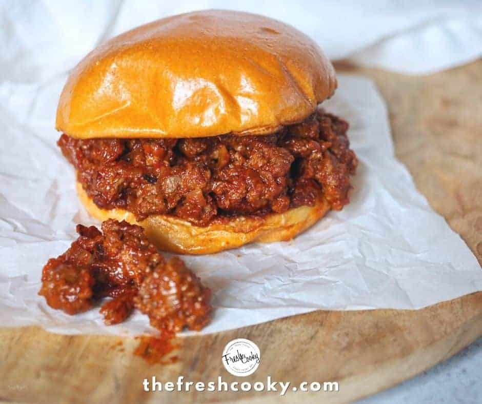 Image of a sloppy joe burger on white parchment paper, with some of the sloppy joe mixture fallen out. All on a wooden charger