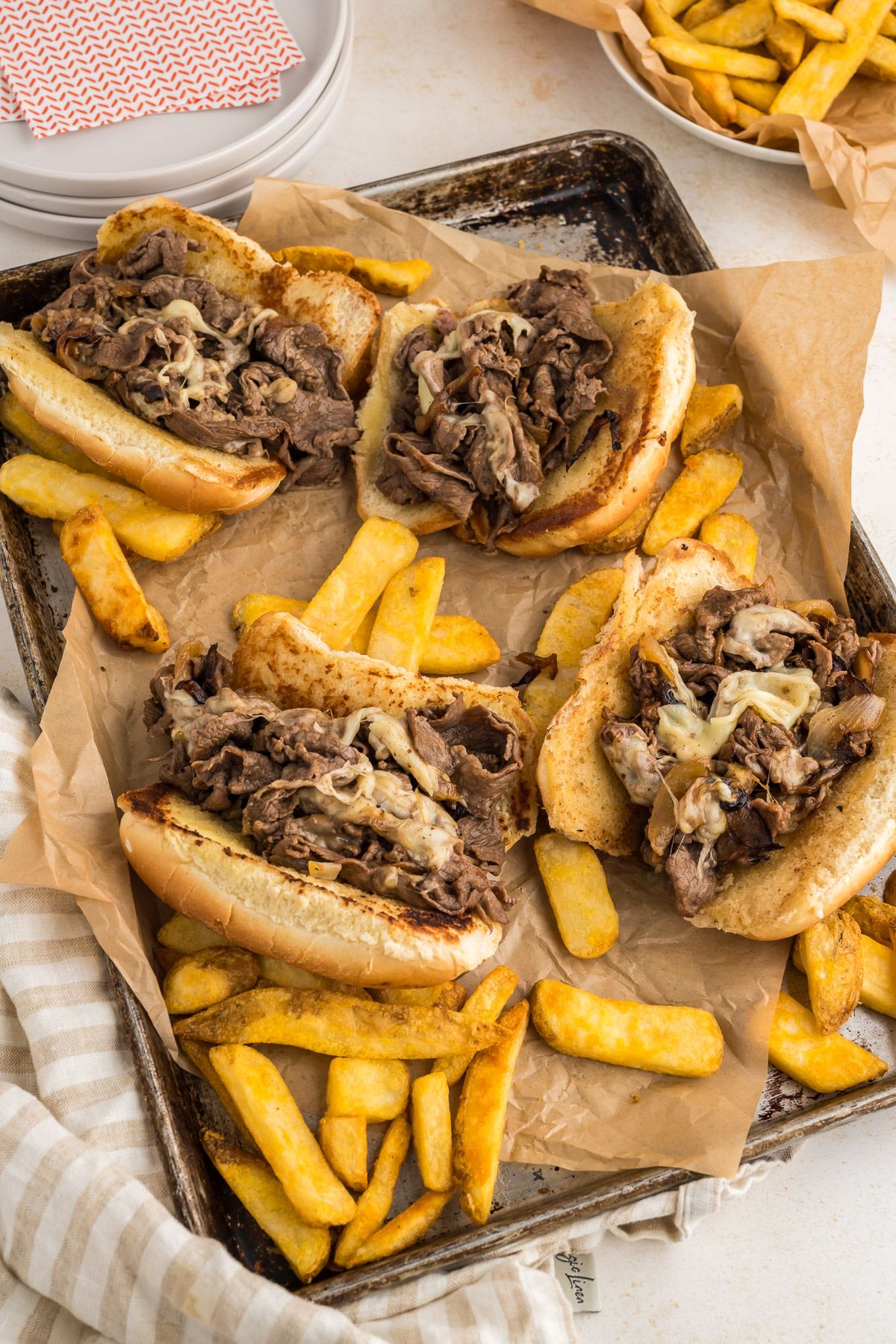 Pan filled with 4 Philly Cheese steak sandwiches with french fries sprinkled around.