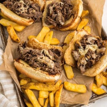 Pan filled with 4 Philly Cheese steak sandwiches with french fries sprinkled around.