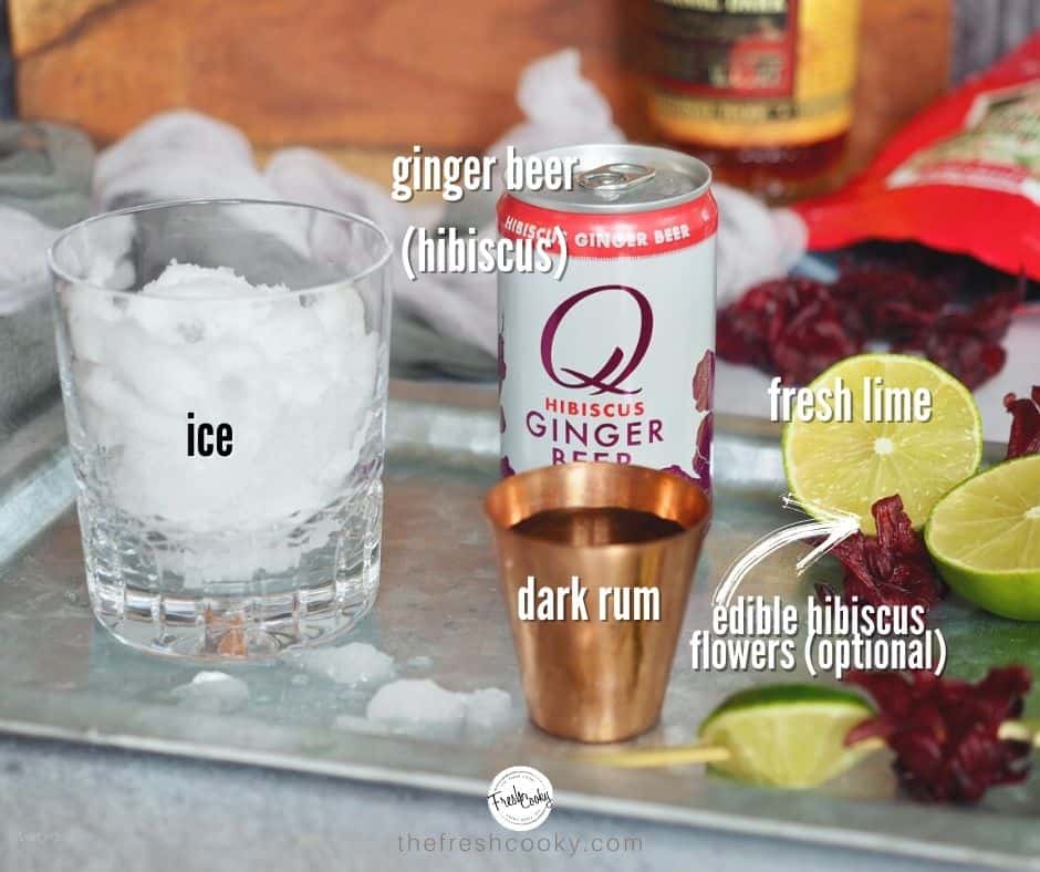 Labeled ingredients for dark and stormy cocktail. left to right, cocktail glass, can of hibiscus ginger beer, limes cut in half, with hibiscus flowers.