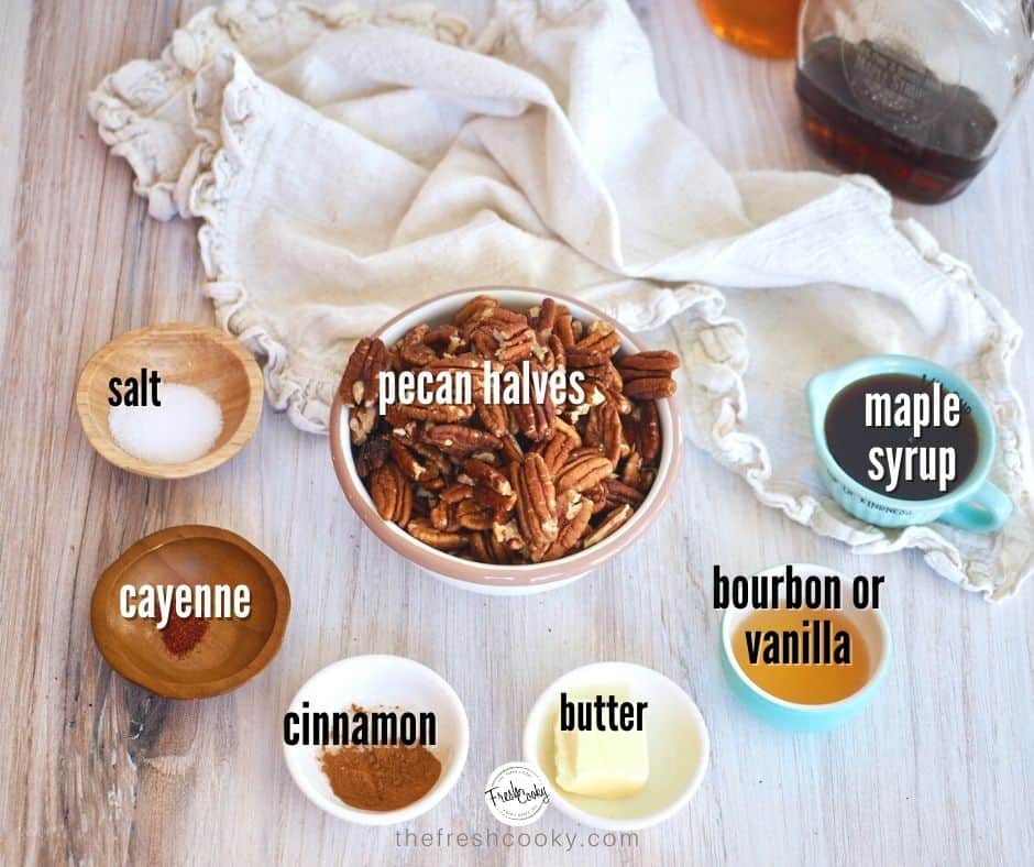 Left to right, ingredient image for Maple Bourbon Pecans. Salt, cayenne pepper, cinnamon, butter, bourbon, maple syrup and pecans, on wood with neutral colored napkin and bottles of maple syrup and bourbon in background.