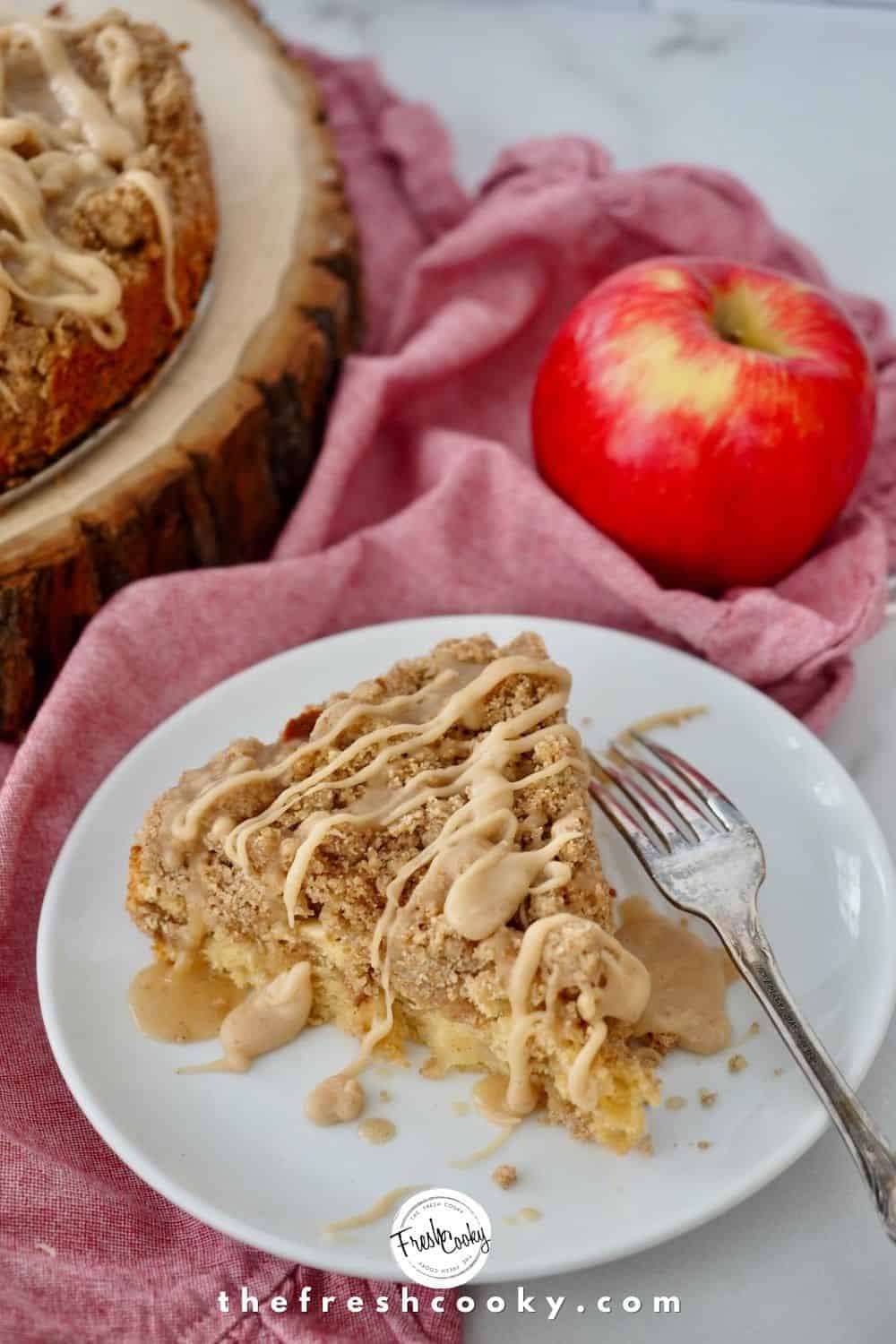 Top down shot of slice of apple crumb cake with fork laying on a white plate atop a pink napkin with a red apple and wooden cake plate in background.