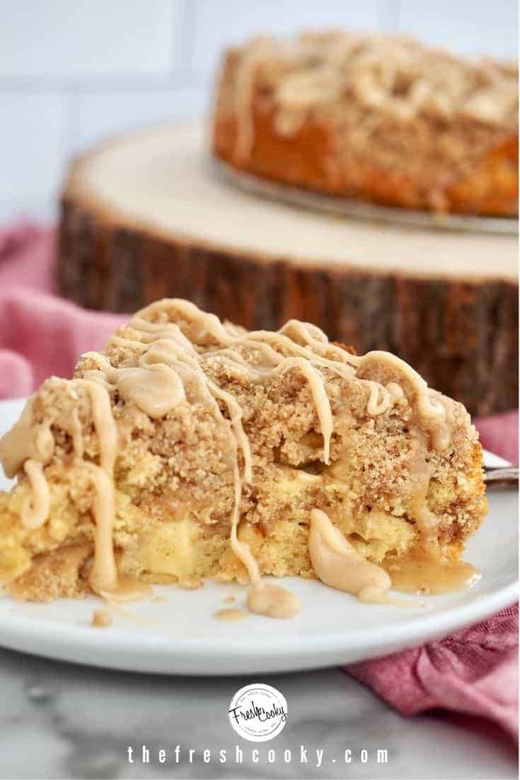 Wedge of apple crumb cake on a white plate sitting on a pink napkin with a wooden cake plate and the rest of the apple crumb cake in the background.