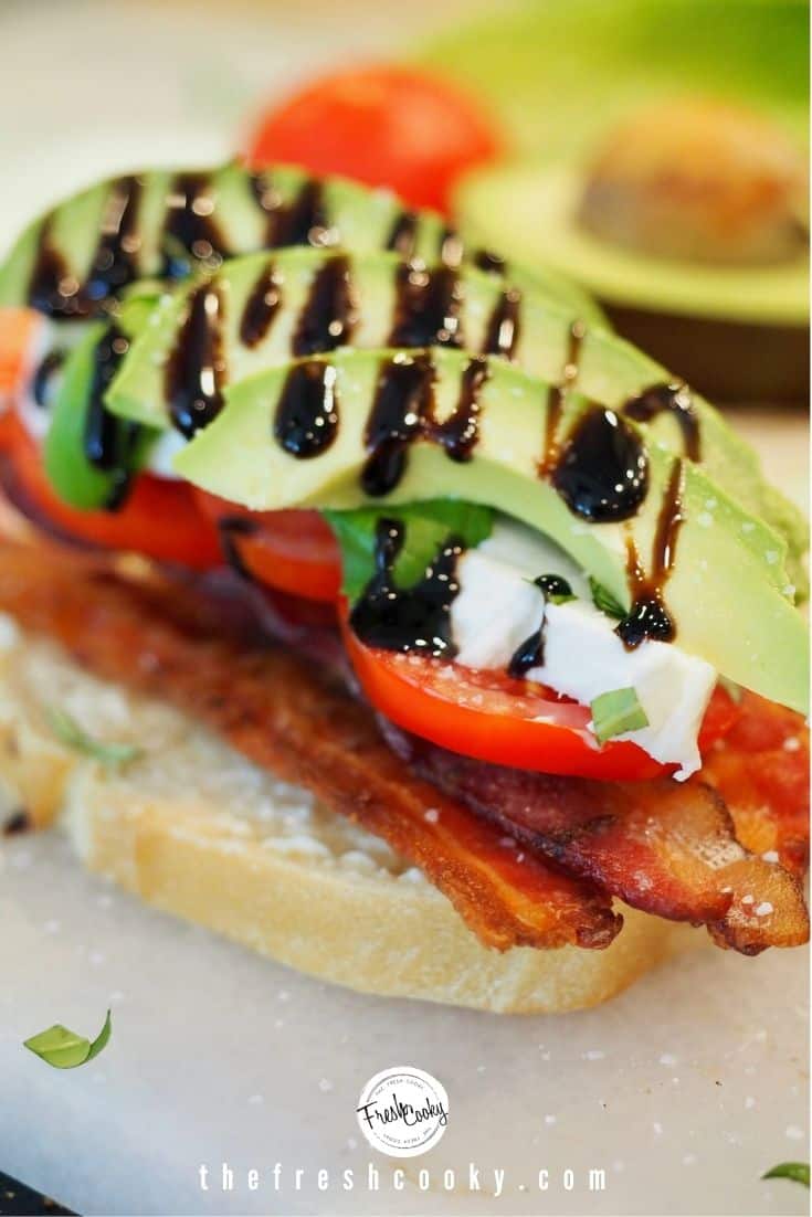 sourdough toast, with mayo, bacon, tomatoes, mozzarella cheese, basil, and avocado, drizzled with balsamic glaze, avocado and tomato halves in background. 