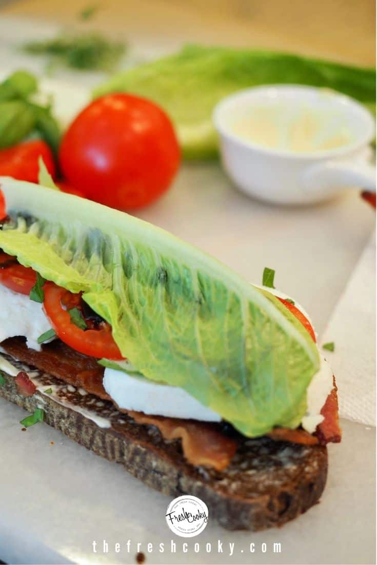 Bacon lettuce and tomato sandwich with added sliced mozzarella, basil and balsamic, topped with a leaf of Romaine lettuce, background has tomato and small bowl of mayo.