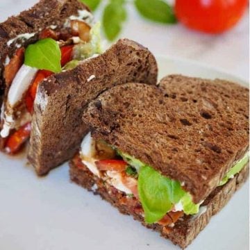 Caprese BLT Sandwich on white plate, cut in half with one half standing on side, bacon lettuce, tomato, mozzarella, basil and balsamic glaze, with pink napkin and red tomato in background.