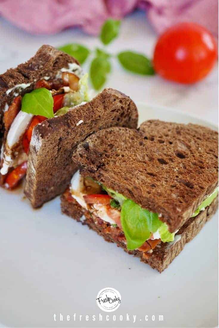 Caprese BLT Sandwich on white plate, cut in half with one half standing on side, bacon lettuce, tomato, mozzarella, basil and balsamic glaze, with pink napkin and red tomato in background.