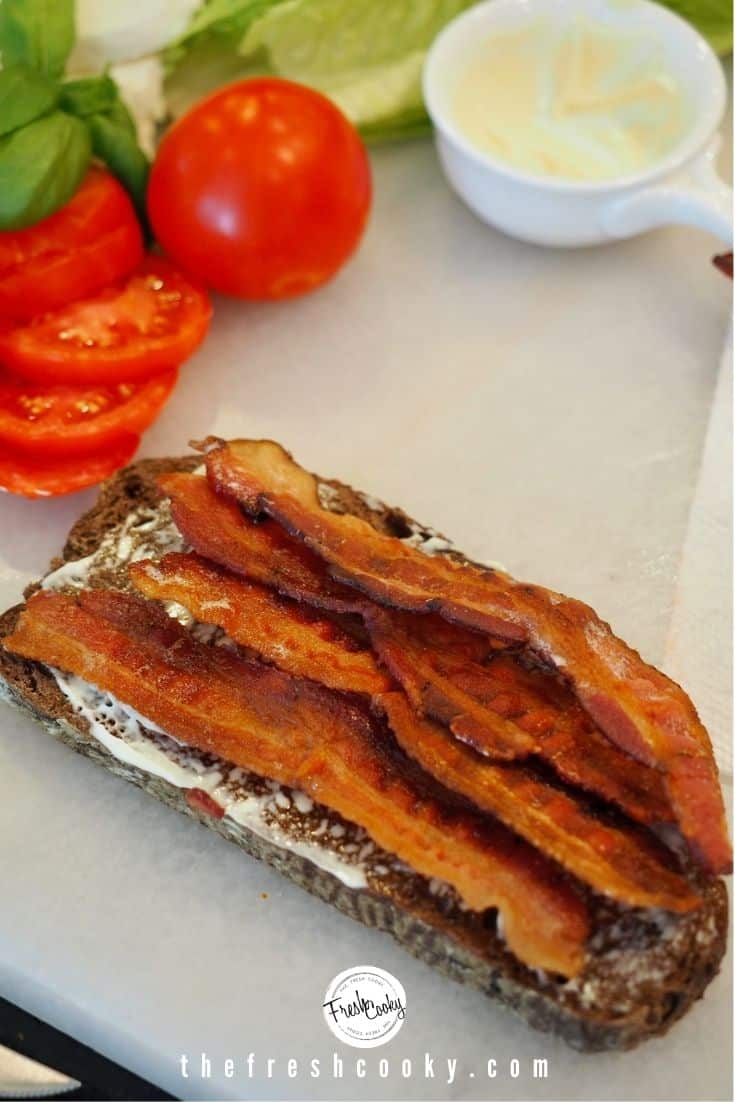 4 slices of crisp bacon on a slice of pumpernickel toast with mayo, slices of tomatoes and basil in background.