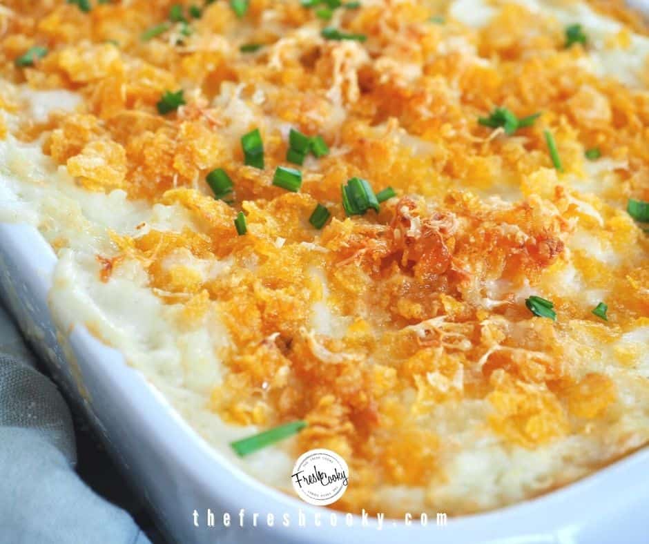 Image of cheesy funeral potatoes in white baking dish, close up shot with crunchy buttery corn flake topping, bubbling cheese and chopped chives.