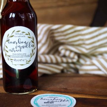 Horizontal image of bottle filled with bourbon vanilla extract with cute label, striped tea towel in background and another label design laying on a cutting board.