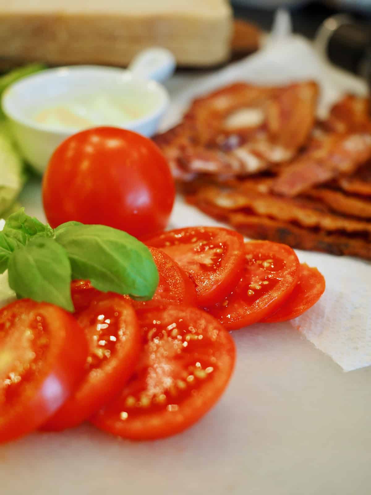 Sliced juicy tomatoes on a cutting board, with a whole tomato behind, fresh basil leaves and in the background crisp bacon and a small white bowl of mayonnaise.