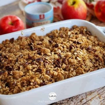 Horizontal image of a white casserole dish filled with baked apple crisp, red apples in the background