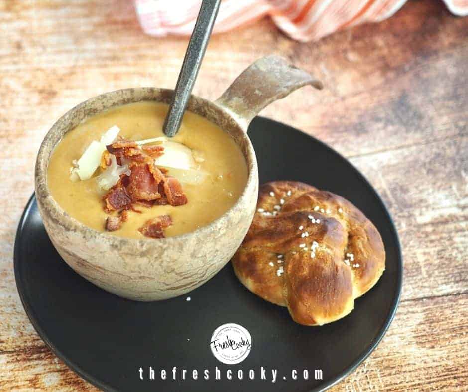 Horizontal image for beer cheese soup with bowl of soup in wooden bowl with spoon on a black plate with soft pretzel beside sitting on a rustic wood surface.